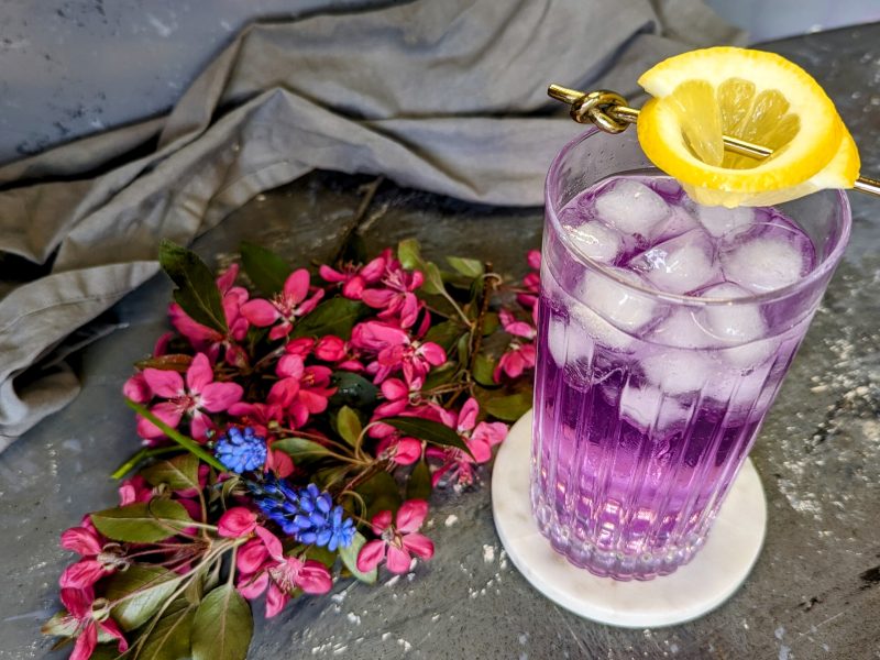 Ein Glas mit dem Butterfly Gin Tonic auf grauem Steintisch mit weißem Untersetzer und roten sowie blauen Blüten.