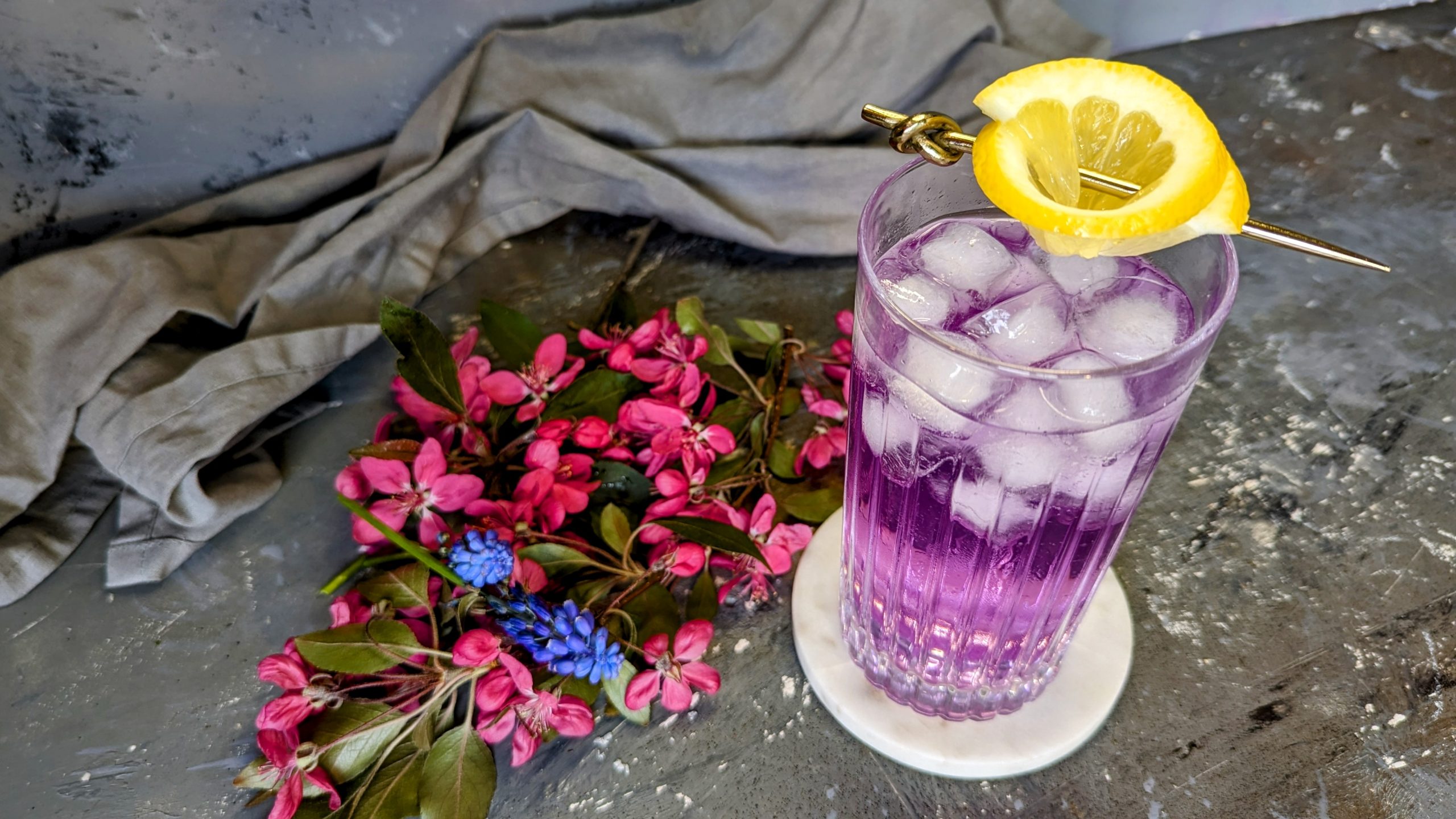 Ein Glas mit dem Butterfly Gin Tonic auf grauem Steintisch mit weißem Untersetzer und roten sowie blauen Blüten.