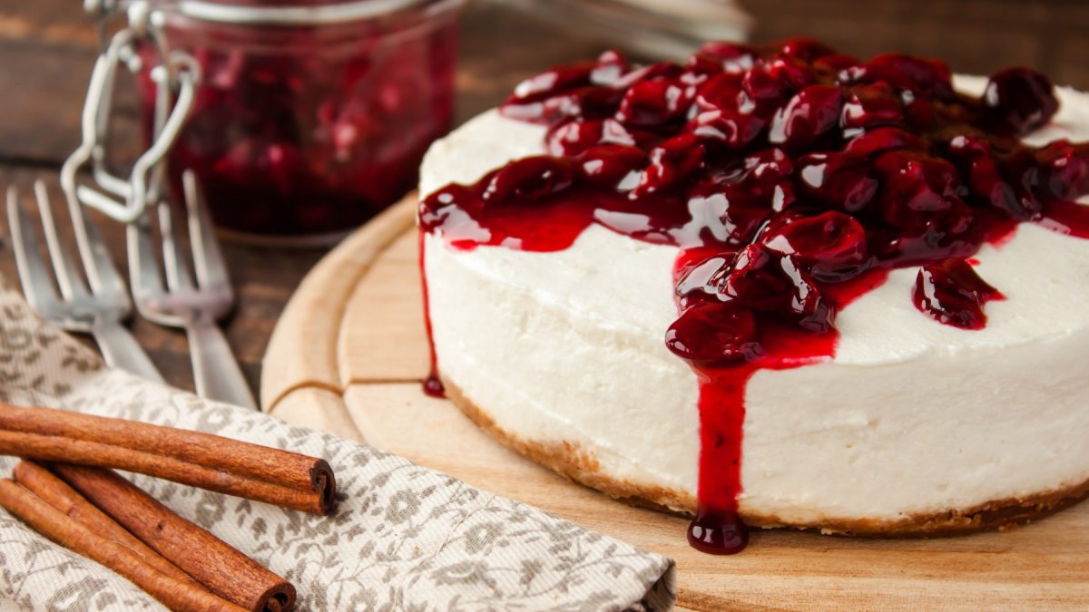 Ein cremiger Quarkkuchen mit Kirschen und Zimtstangen liegt auf einem Holzbrett neben einer Serviette mit Gabeln.