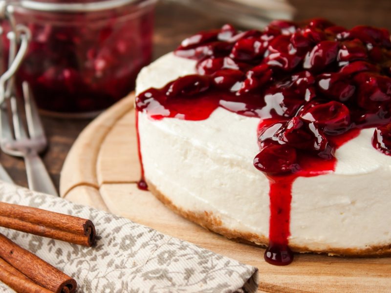 Ein cremiger Quarkkuchen mit Kirschen und Zimtstangen liegt auf einem Holzbrett neben einer Serviette mit Gabeln.