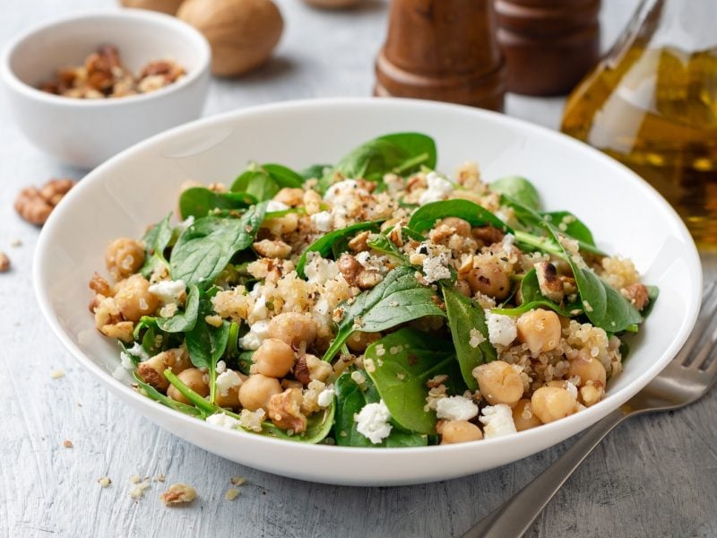 Ein weißer, tiefer Teller mit dem Quinoa-Salat mit Kichererbsen, Feta und Spinat auf einem hellen Holztisch. Im Hintergrund Walnüsse, Salz, Pfeffer und Olivenöl. Alles direkt von leicht schräg oben fotografiert.