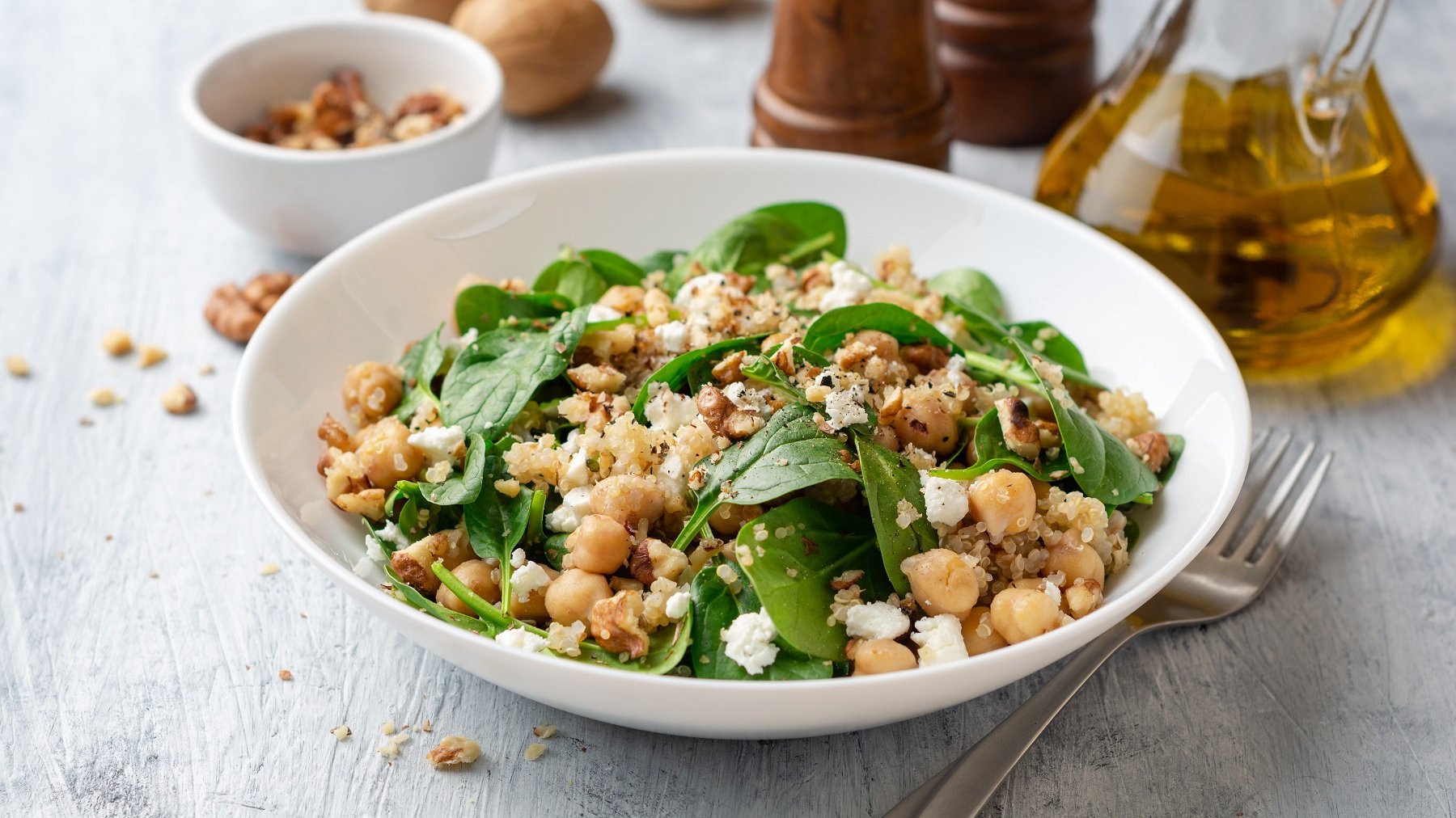 Ein weißer, tiefer Teller mit dem Quinoa-Salat mit Kichererbsen, Feta und Spinat auf einem hellen Holztisch. Im Hintergrund Walnüsse, Salz, Pfeffer und Olivenöl. Alles direkt von leicht schräg oben fotografiert.