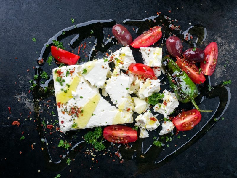 Feta mit Gemüse und Olivenöl von oben fotografiert vor einem dunklen Untergrund.