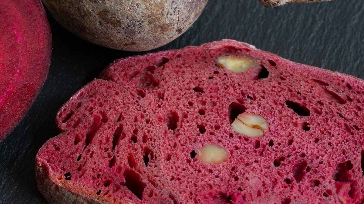 Eine Scheibe vom Rote-Bete-Brot auf einer schwarzen Schiefertafel mit frischer Roter-Bete. Alles direkt von oben und recht nah fotografiert.