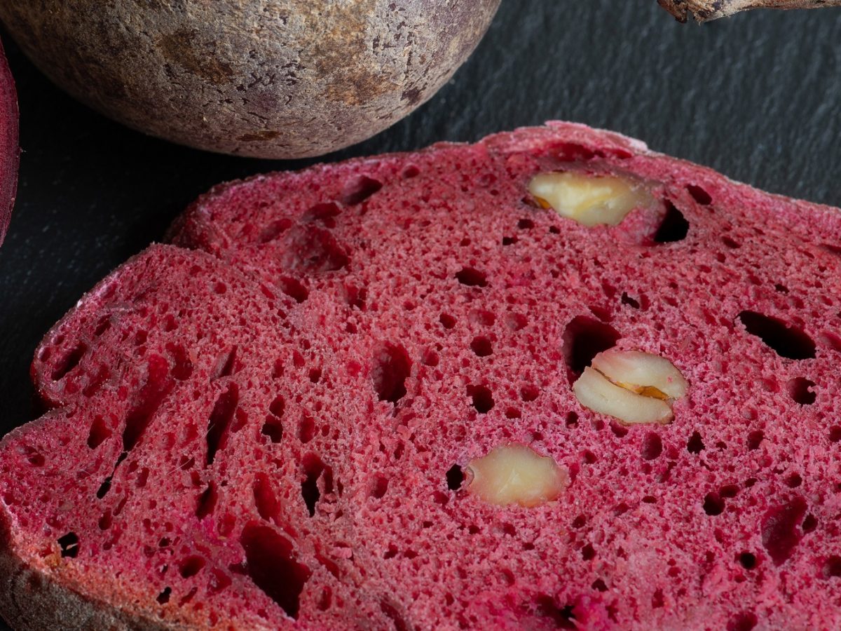 Eine Scheibe vom Rote-Bete-Brot auf einer schwarzen Schiefertafel mit frischer Roter-Bete. Alles direkt von oben und recht nah fotografiert.