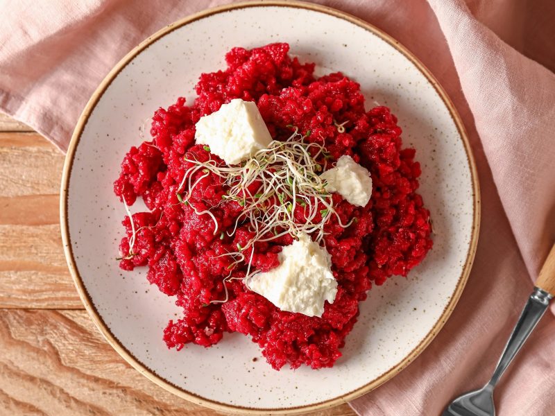 Ein beiger Teller mit dem Risotto auf einem hellen Holztisch mit rosa Tischdecke. Alles direkt von oben fotografiert.