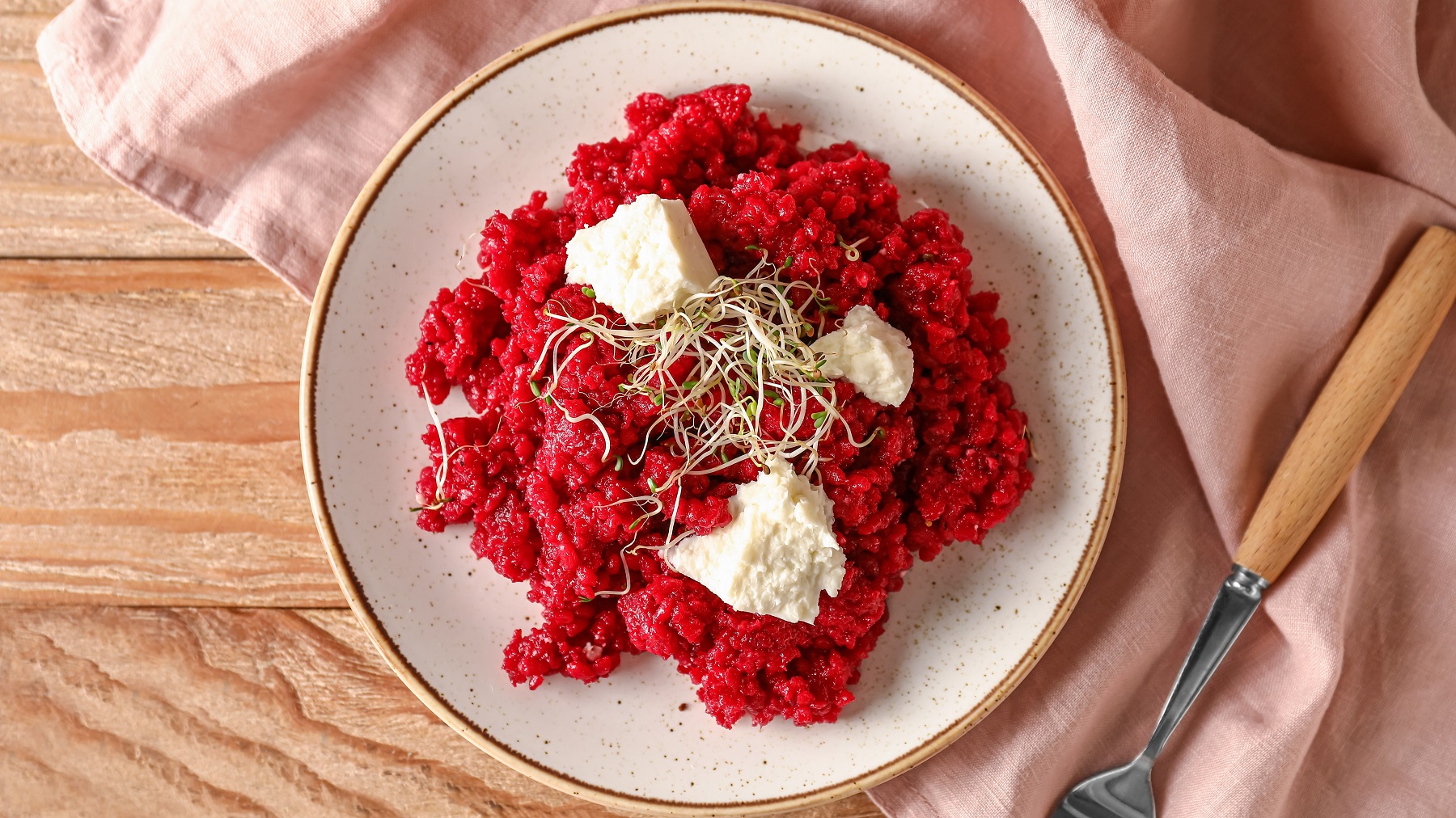 Ein beiger Teller mit dem Risotto auf einem hellen Holztisch mit rosa Tischdecke. Alles direkt von oben fotografiert.