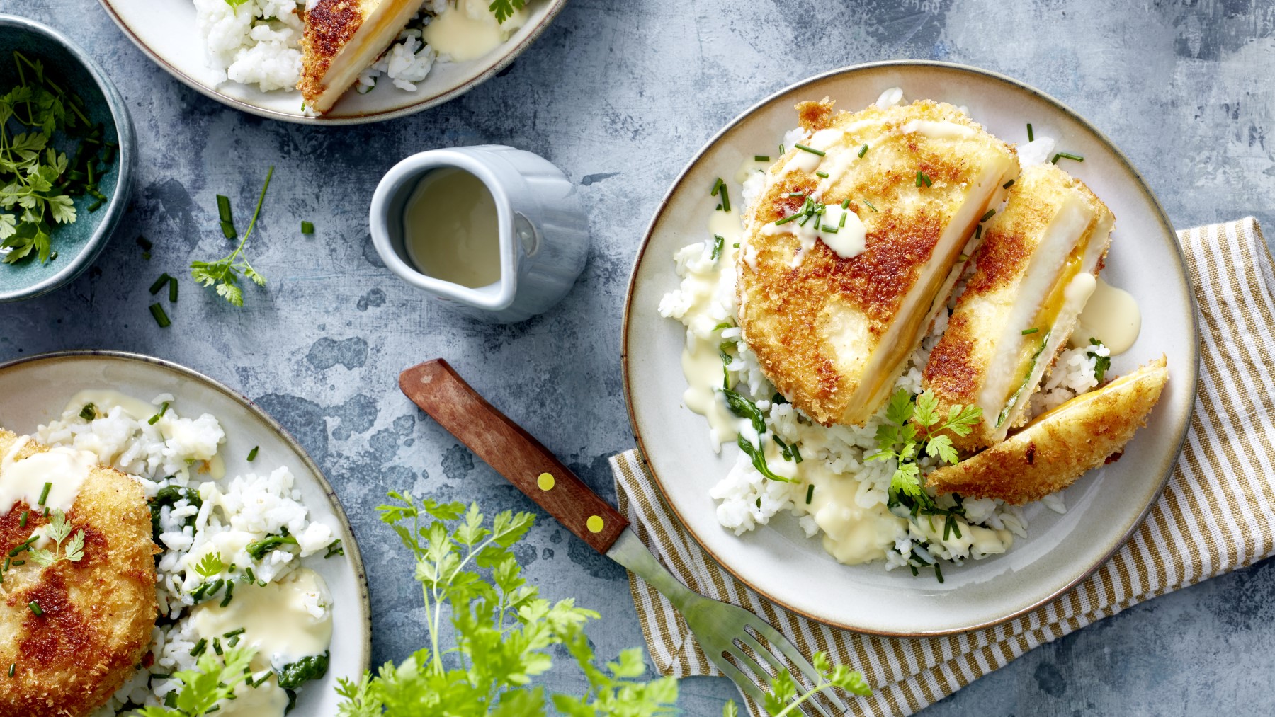 Teilweise Ansicht von drei Tellern mit Sellerie-Cordon-bleu, Reis, heller Sauce und Kräutern.