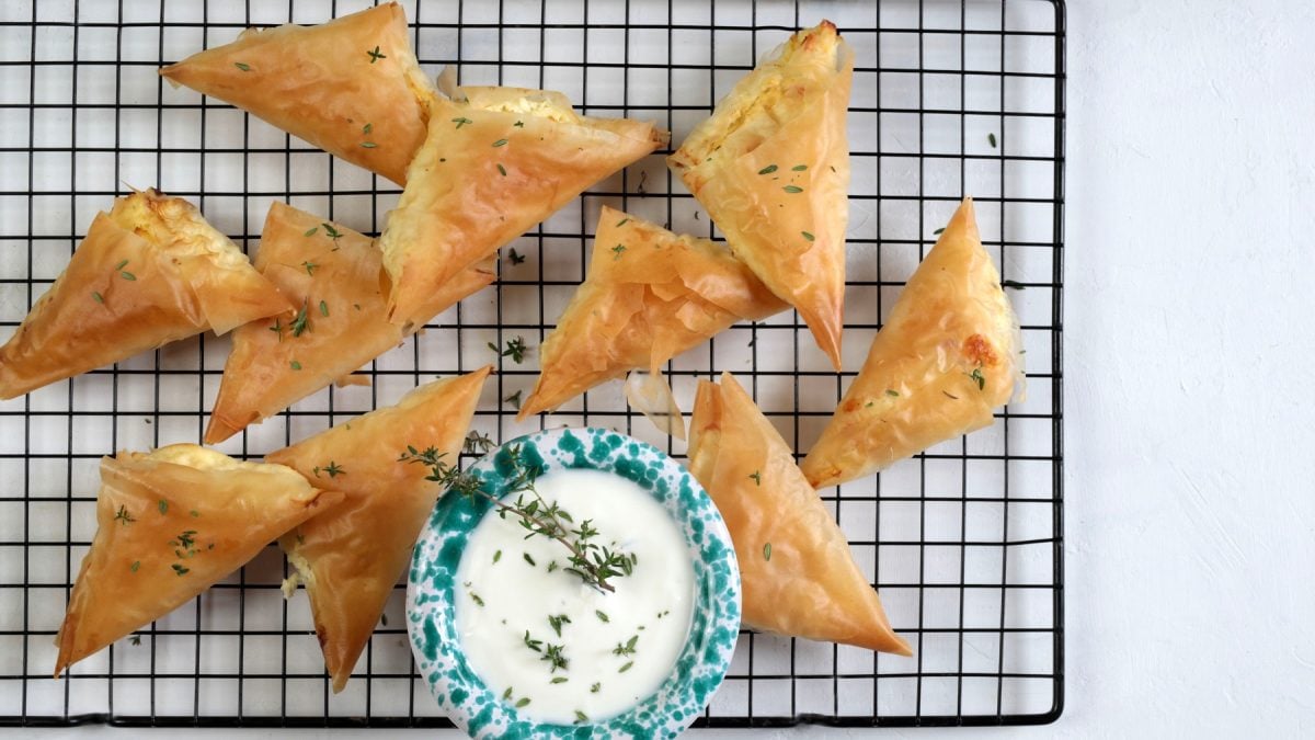 Frisch gebackene, dreieckige Tiropitakia auf einem schwarzen Gitter, garniert mit Kräutern und serviert mit einem Joghurt-Dip.