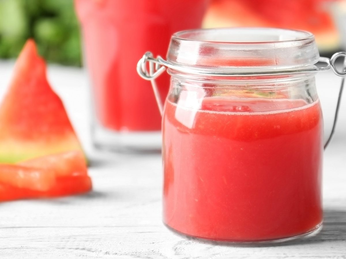 Ein Glas mit der roten Marmelade auf einem weißen Tisch mit frischer Melone im Hintergrund.