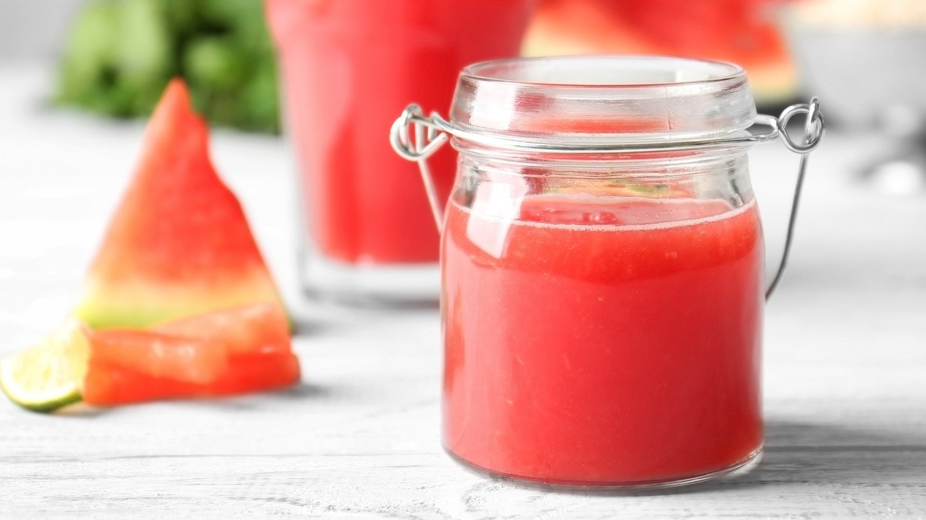 Ein Glas mit der roten Marmelade auf einem weißen Tisch mit frischer Melone im Hintergrund.