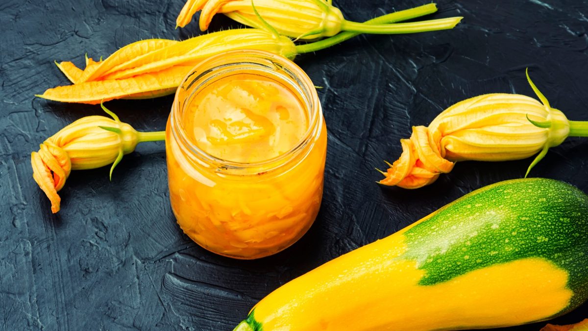 Ein Glas mit dem Zgelben Zucchini-Cutney und gelber Zucchini sowie Blüten auf einem schwarzen Tisch.