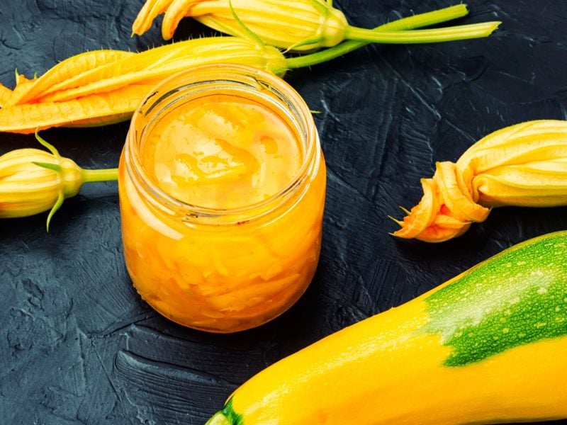 Ein Glas mit dem Zgelben Zucchini-Cutney und gelber Zucchini sowie Blüten auf einem schwarzen Tisch.