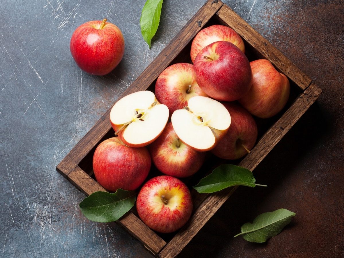 Eine Holzkiste voller roter Äpfel und ein halbierter Apfel liegen auf einer rustikal zerkratzten grauen Steinfläche.