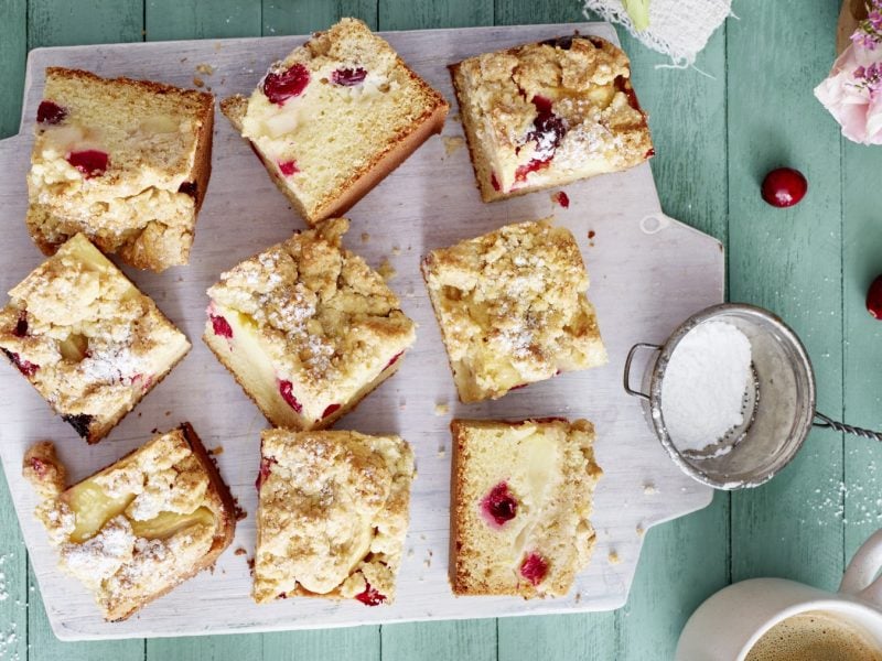 Apfel-Cranberry-Streuselkuchen auf einem weißen Brett auf grünem Hintergrund. Links Cranberrys und rechts Puderzucker und Blumen.