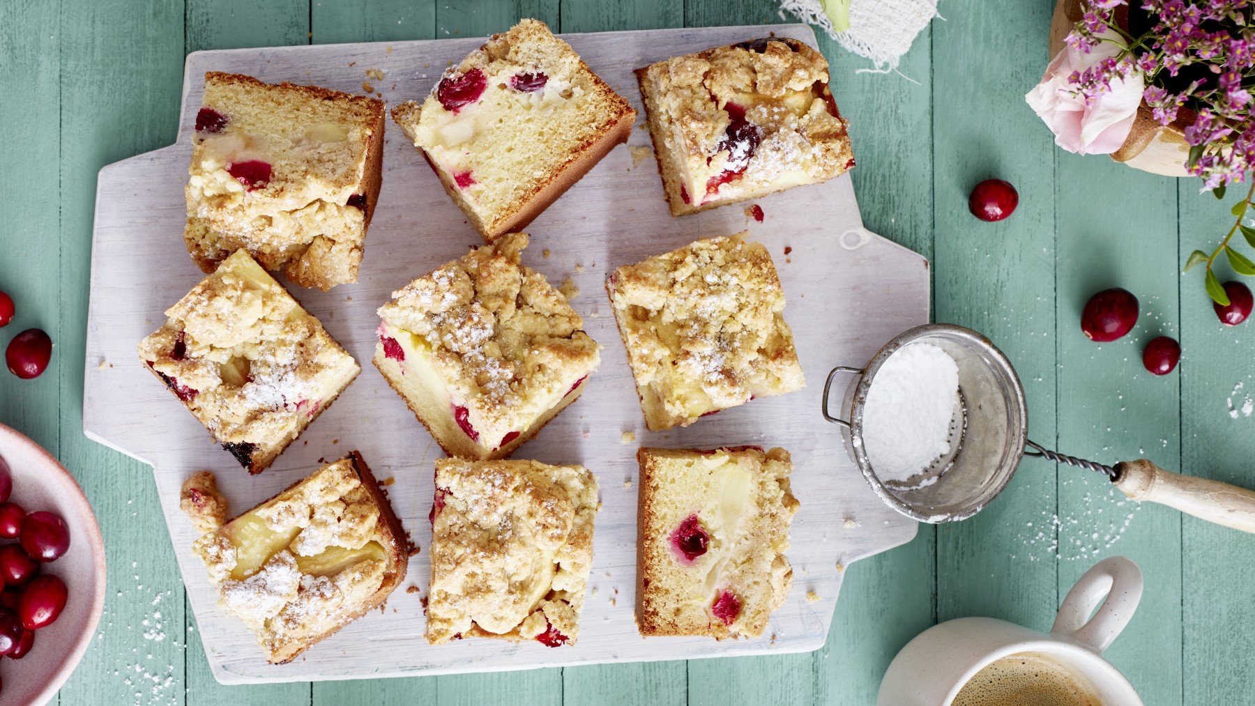 Apfel-Cranberry-Streuselkuchen auf einem weißen Brett auf grünem Hintergrund. Links Cranberrys und rechts Puderzucker und Blumen.