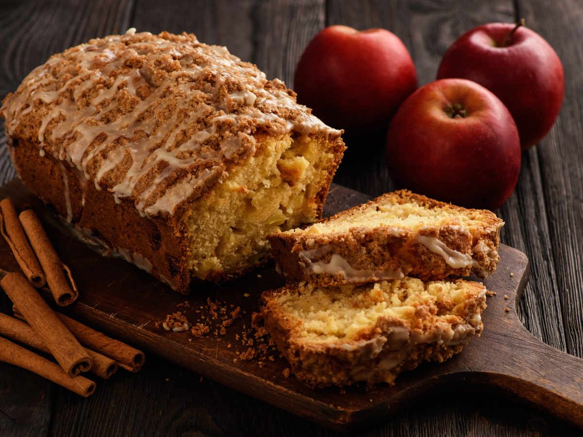 Apfel-Zimt-Kuchen auf einem Holzbrett auf hölzernem Hintergrund. Rechts davon Äpfel und links davon Zimtstangen.