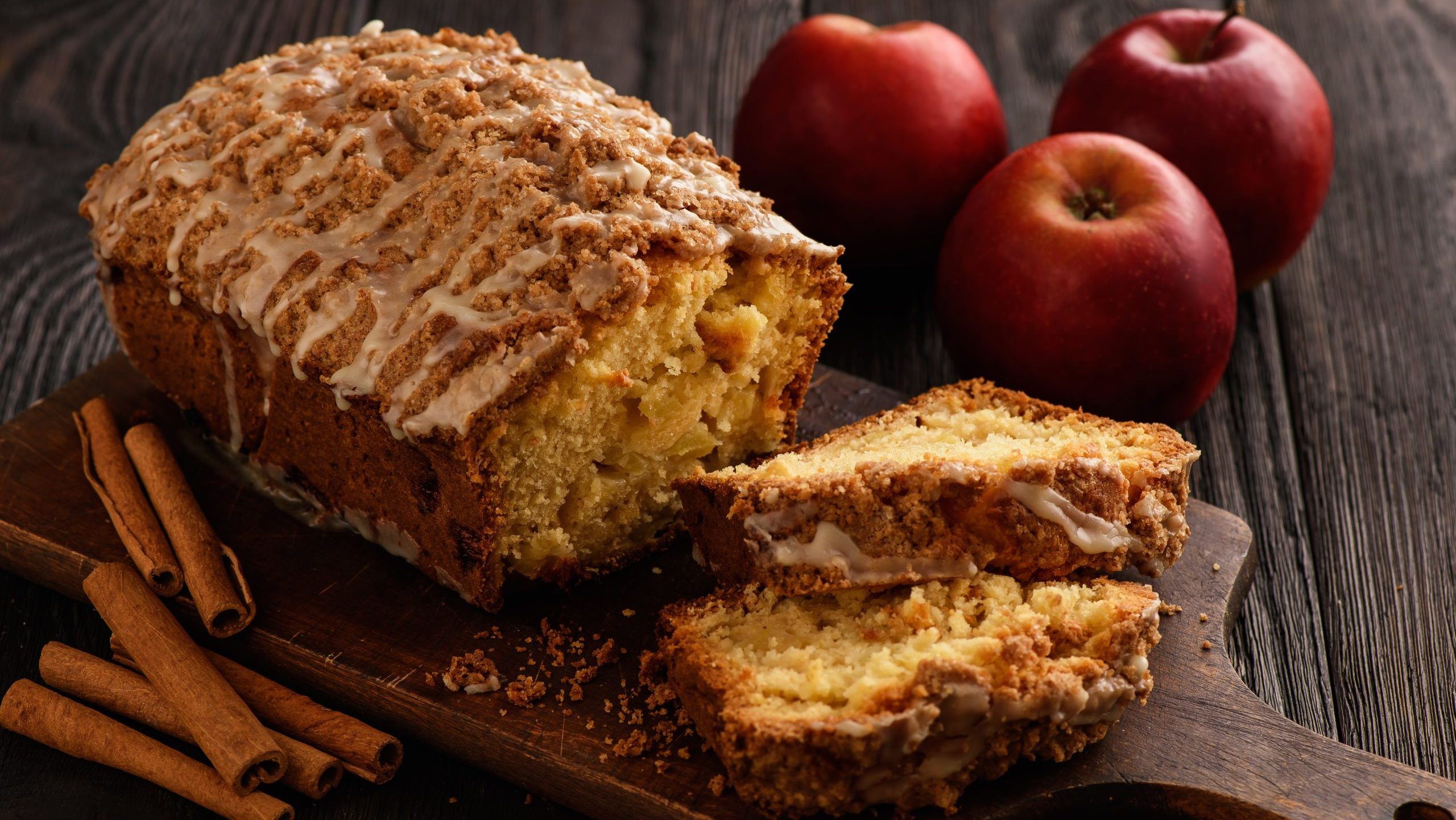 Apfel-Zimt-Kuchen auf einem Holzbrett auf hölzernem Hintergrund. Rechts davon Äpfel und links davon Zimtstangen.