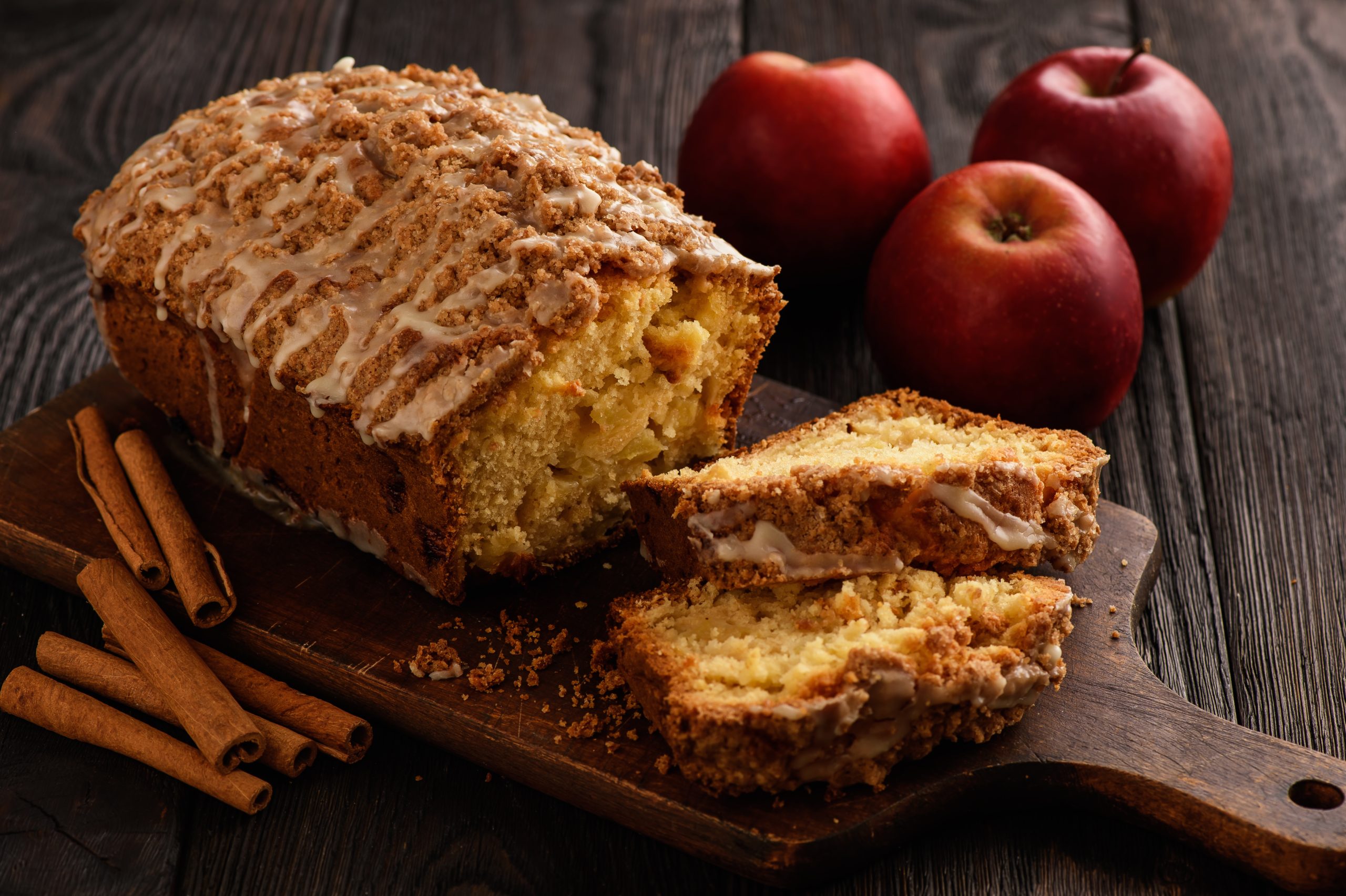 Apfel-Zimt-Kuchen auf einem Holzbrett auf hölzernem Hintergrund. Rechts davon Äpfel und links davon Zimtstangen.