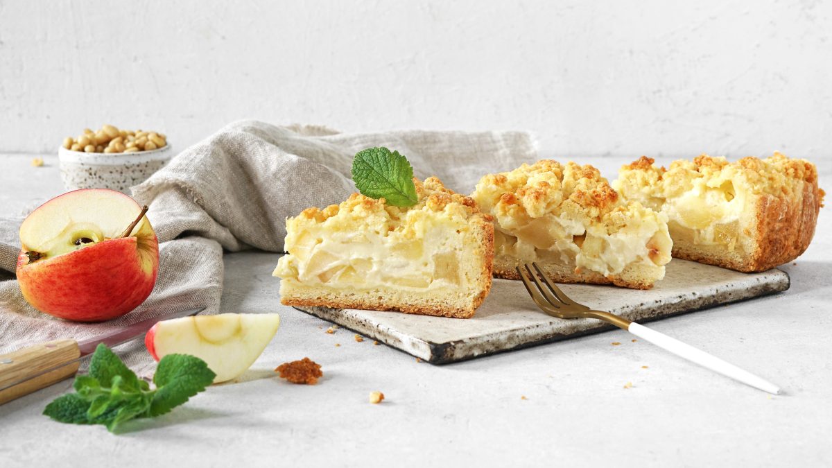 Drei Stücken vom Apfelkuchen mit Pudding auf grauem Untergrund mit hellgrauem Hintergrund. Alles von der Seite fotografiert und daneben frischer Apfel und grüne Minze.