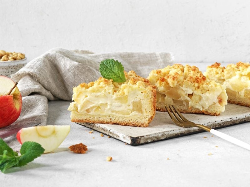 Drei Stücken vom Apfelkuchen mit Pudding auf grauem Untergrund mit hellgrauem Hintergrund. Alles von der Seite fotografiert und daneben frischer Apfel und grüne Minze.