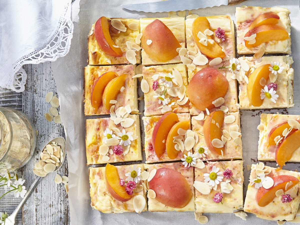 Aprikosen-Kuchen vom Blech auf Backpapier auf hölzernem Hintergrund. Links davon ein Löffel, Mandelblättchen und essbare Blüten.