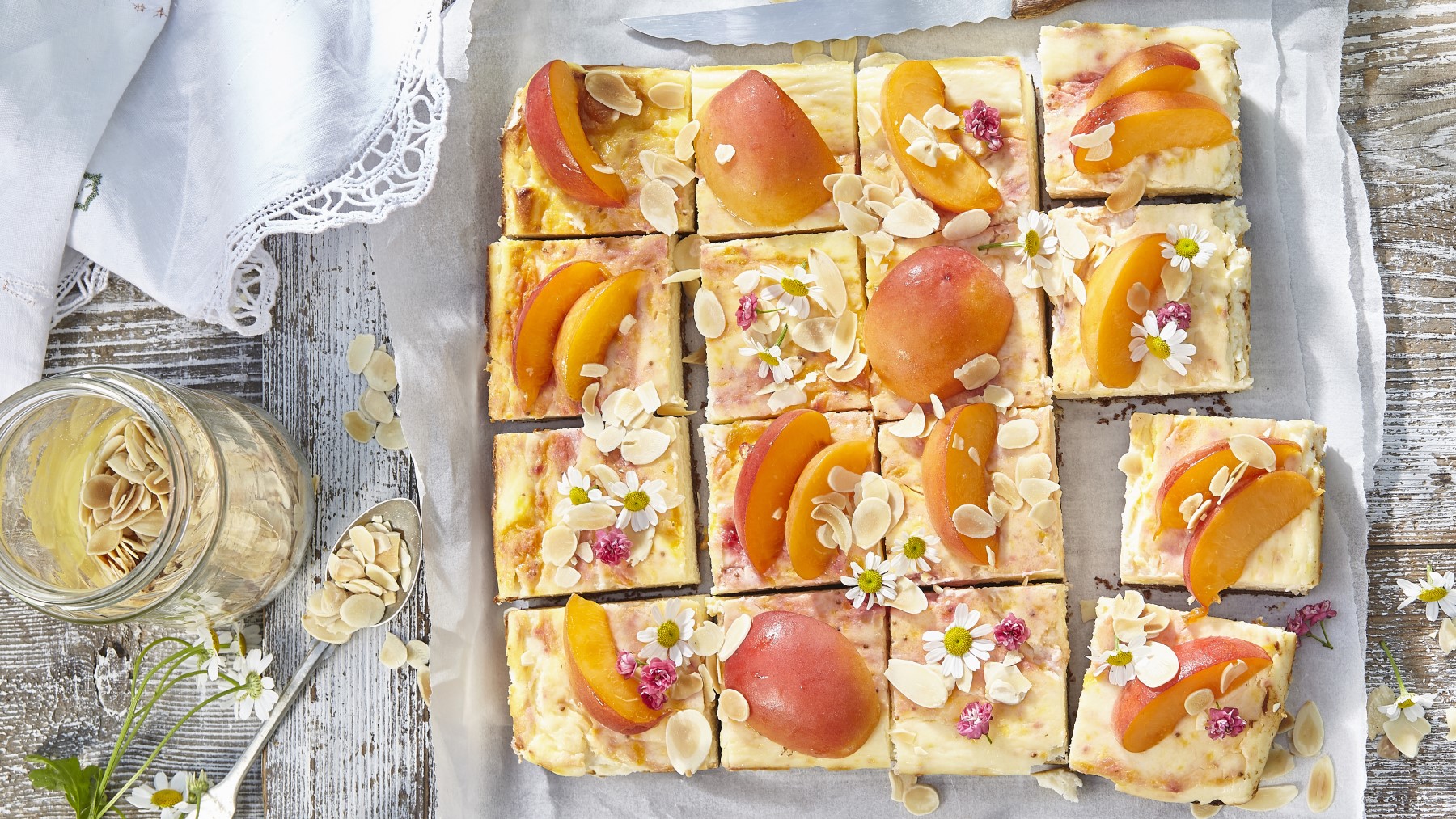 Aprikosen-Kuchen vom Blech auf Backpapier auf hölzernem Hintergrund. Links davon ein Löffel, Mandelblättchen und essbare Blüten.