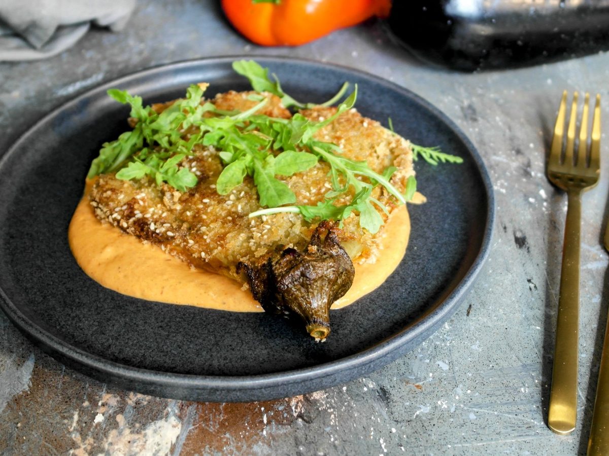 Ein schwarzer Teller mit der knusprigen Aubergine und rötlicher Sauce auf grauem Steinuntergrund mit goldenem Besteck von schräg oben fotografiert.