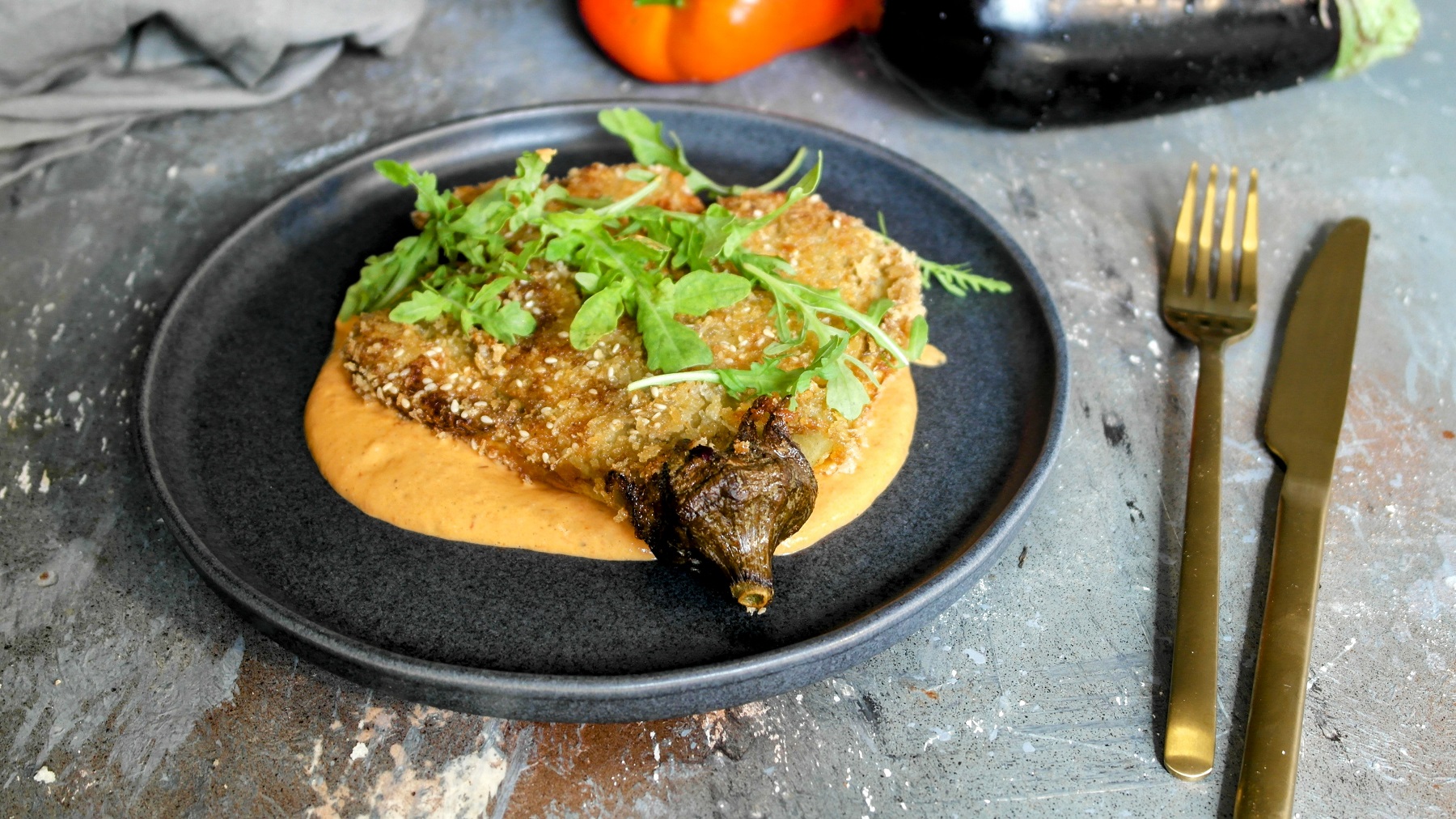 Ein schwarzer Teller mit der knusprigen Aubergine und rötlicher Sauce auf grauem Steinuntergrund mit goldenem Besteck von schräg oben fotografiert.