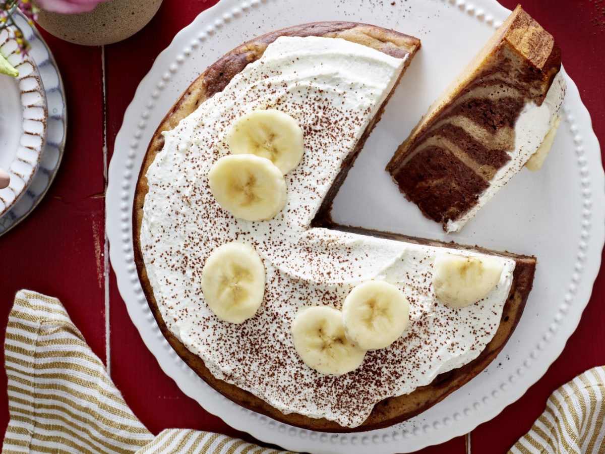 Ein Zebra-Bananen-Kuchen auf einem weißen Teller auf rotem Hintergrund. Links eine Tasse Kaffee und unten ein Geschirrtuch.