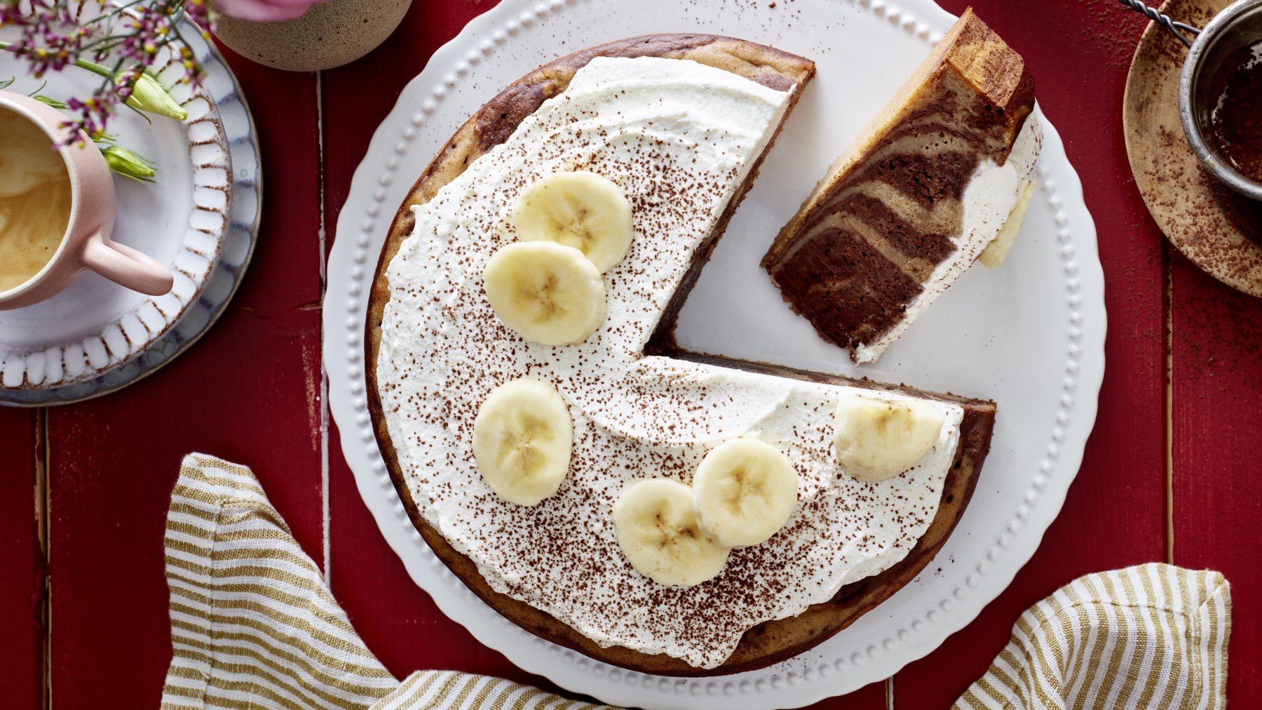 Ein Zebra-Bananen-Kuchen auf einem weißen Teller auf rotem Hintergrund. Links eine Tasse Kaffee und unten ein Geschirrtuch.