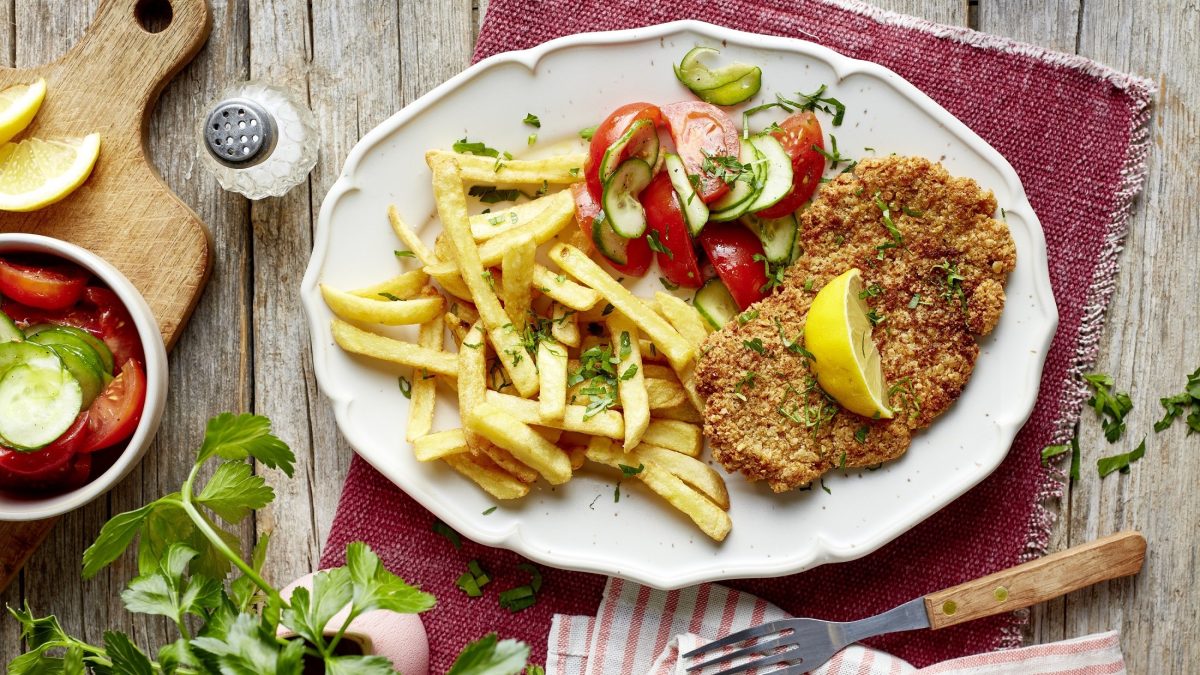 Draufsicht: Ein Haferflocken-Schnitzel mit Salat auf einem weißen, ovalen Teller. Der steht auf einem rustikalen Holztisch.