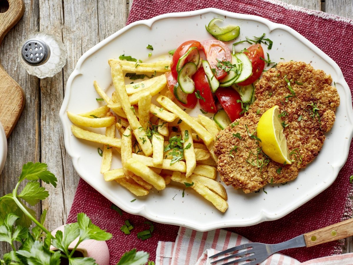 Draufsicht: Ein Haferflocken-Schnitzel mit Salat auf einem weißen, ovalen Teller. Der steht auf einem rustikalen Holztisch.