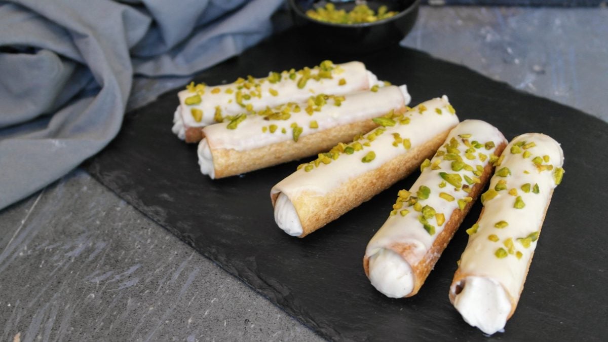 Fünf Cannoli auf einer schwarzen Schieferplatte und grauem Steintisch mit grauem Tuch im Hintergrund. Alles von schräg oben fotografiert.