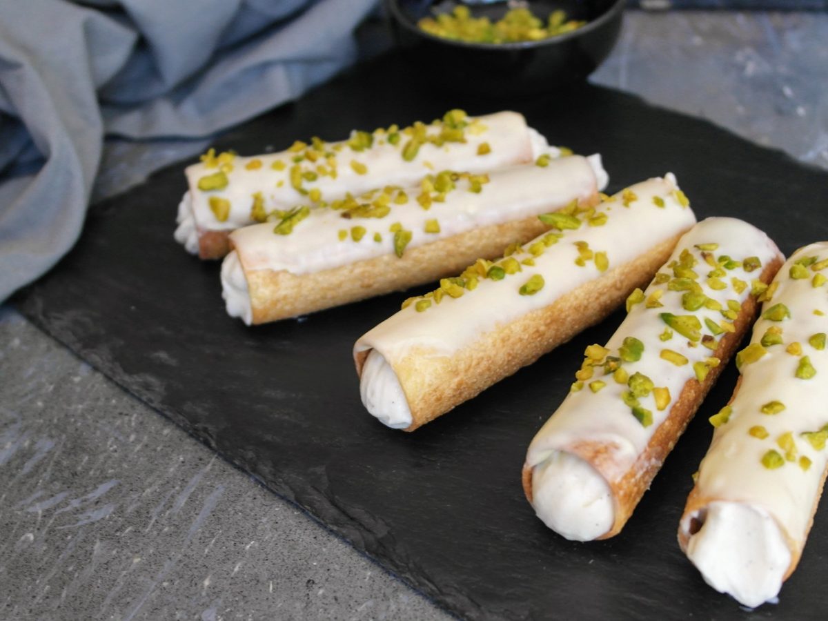 Fünf Cannoli auf einer schwarzen Schieferplatte und grauem Steintisch mit grauem Tuch im Hintergrund. Alles von schräg oben fotografiert.