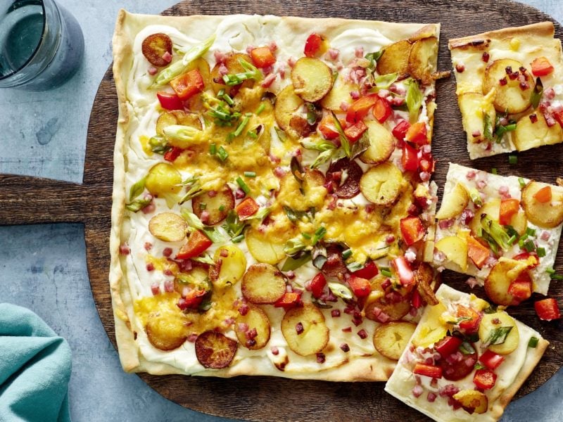 Speck-Flammkuchen auf einem schwarzen Blech auf grauem Hintergrund. Links ein Geschirrtuch, rechts ein Schneidebrett mit Schnittlauch.