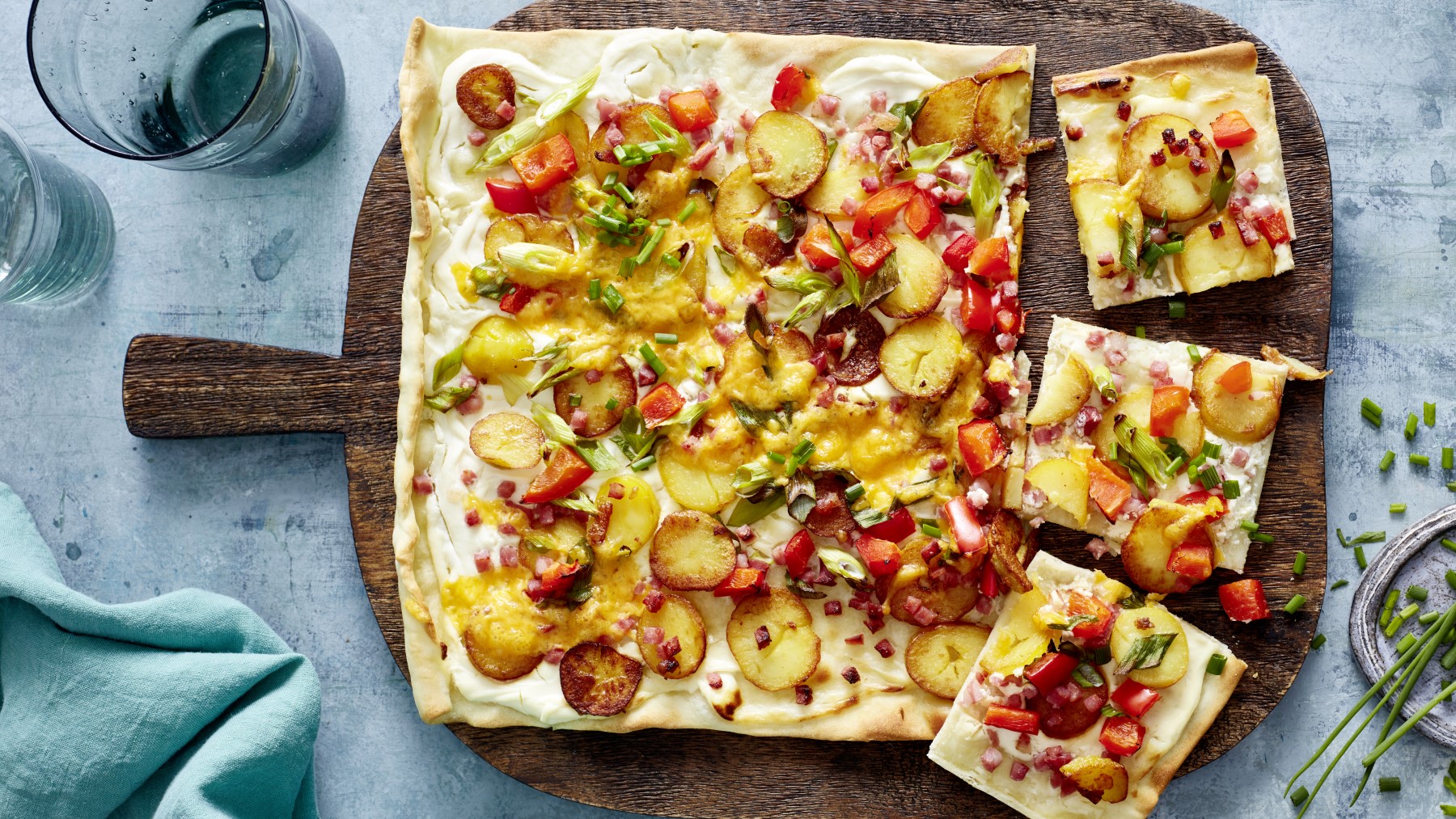 Speck-Flammkuchen auf einem schwarzen Blech auf grauem Hintergrund. Links ein Geschirrtuch, rechts ein Schneidebrett mit Schnittlauch.