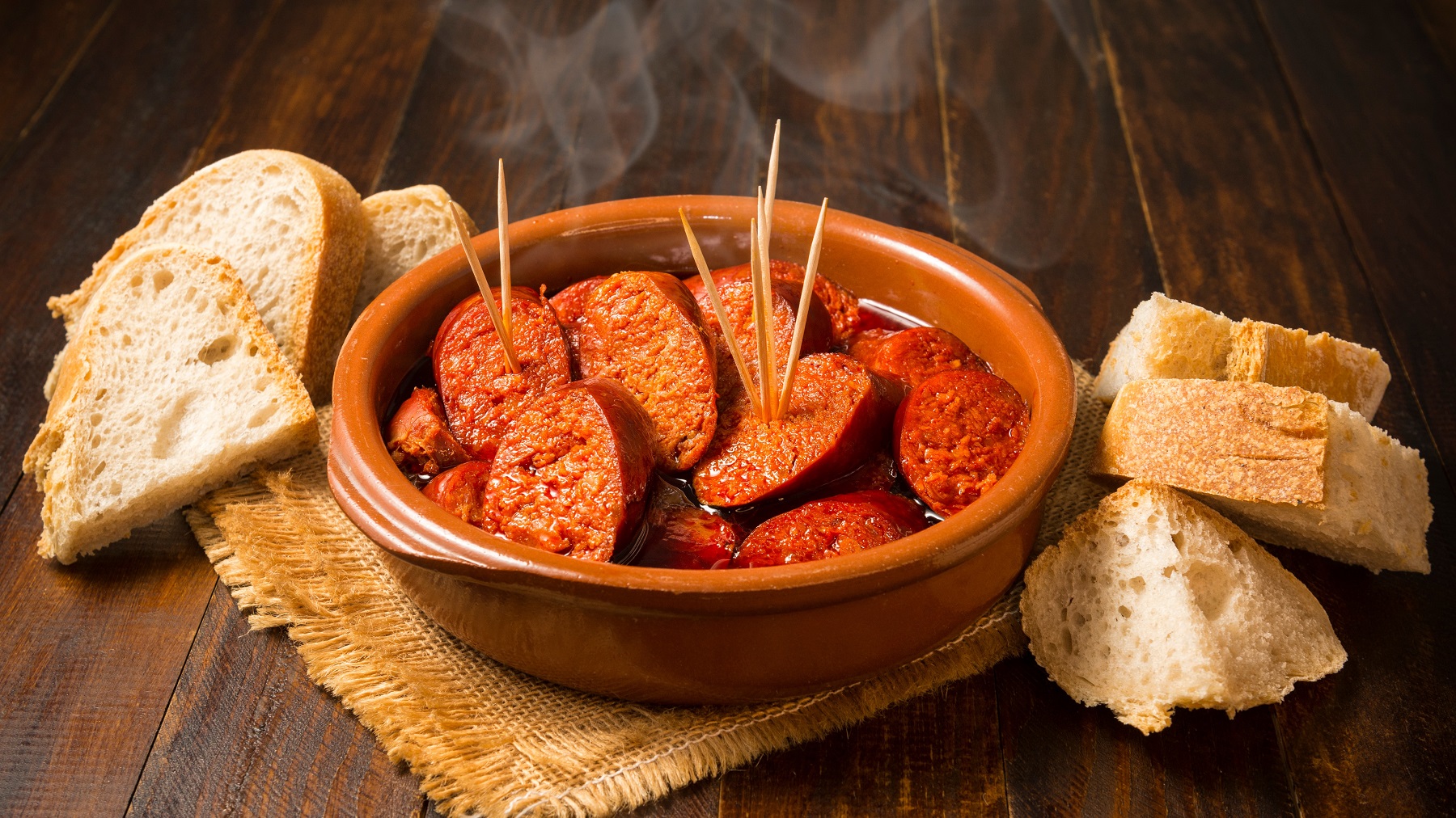 Chorizo a la sidra in einer braunen Tonschale, mit Zahnstochern und Brotstücken.