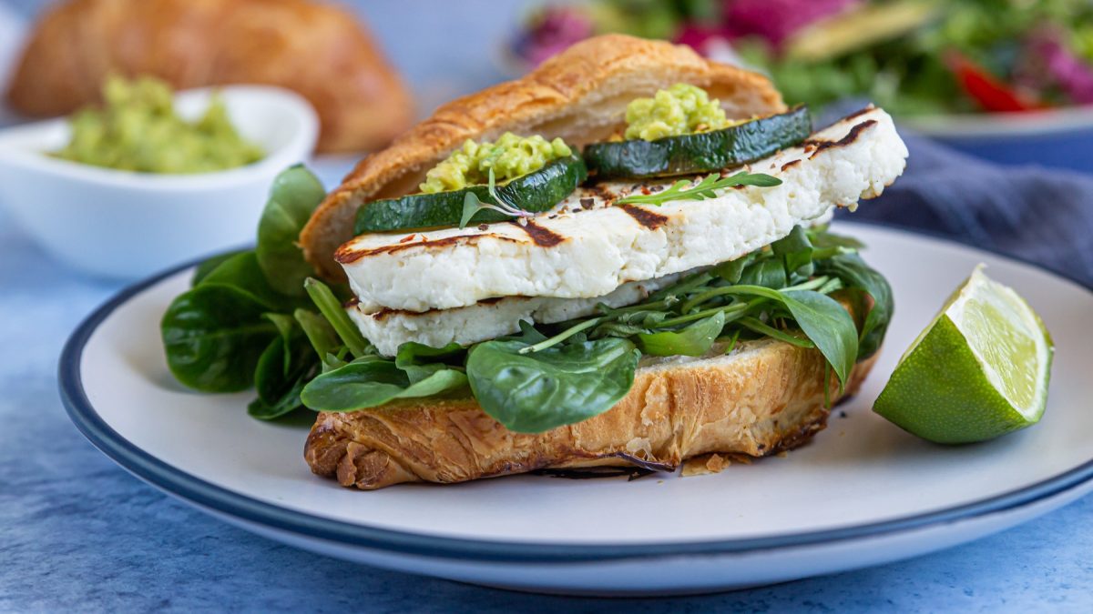 Croissant-Sandwich mit Rucola, Halloumi, Zucchini und pesto, daneben Limettenspalte, auf einem weißen teller.