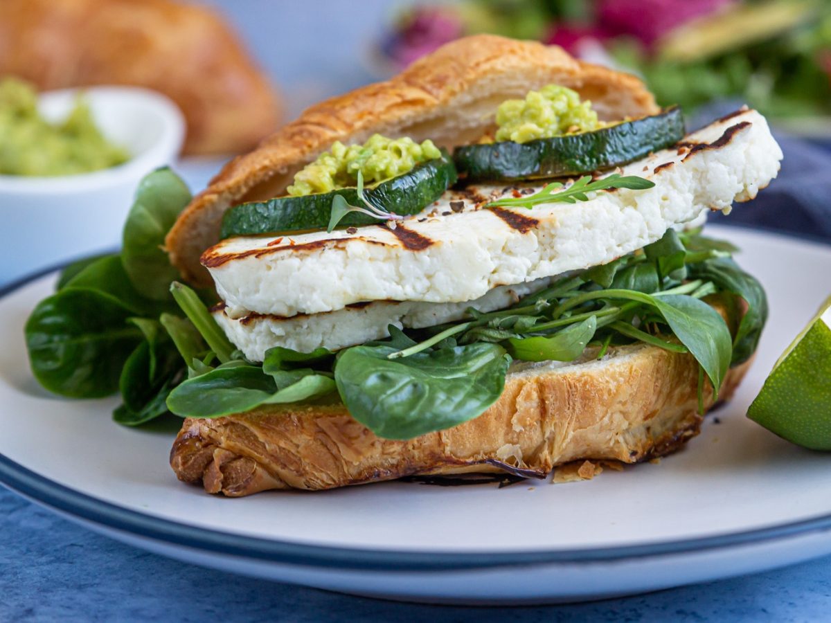 Croissant-Sandwich mit Rucola, Halloumi, Zucchini und pesto, daneben Limettenspalte, auf einem weißen teller.