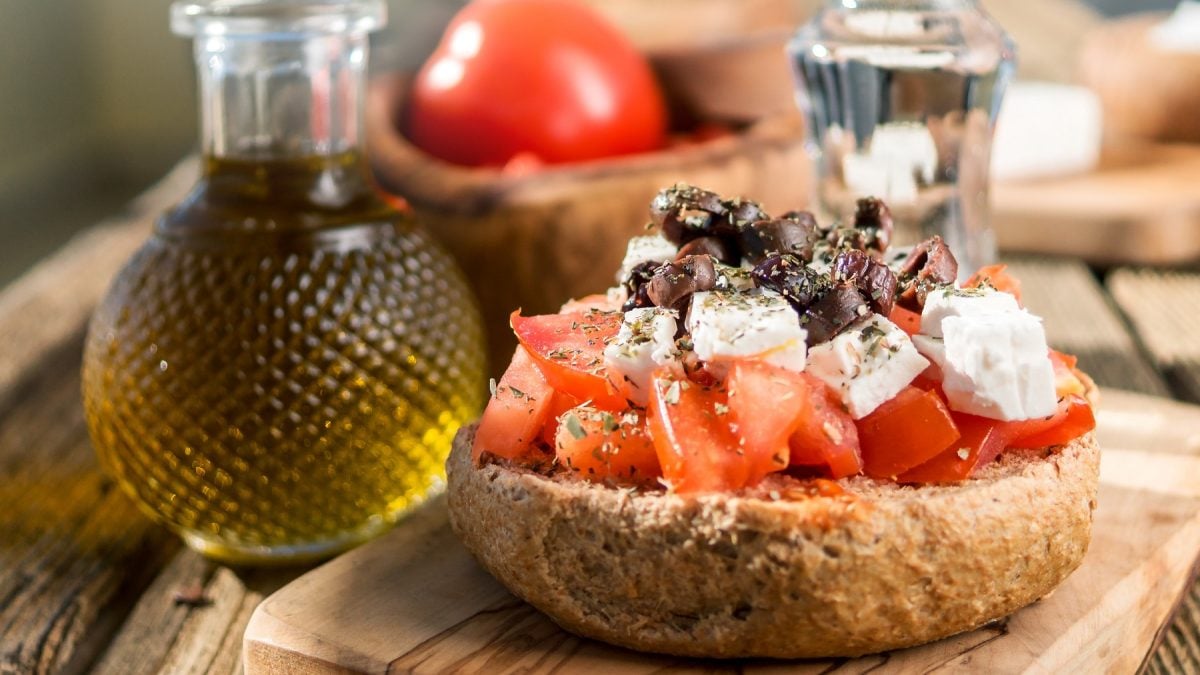 Dakos mit Tomaten, Feta, Oliven und Oregano, daneben eine Flasche mit Olivenöl von vorne fotografiert.