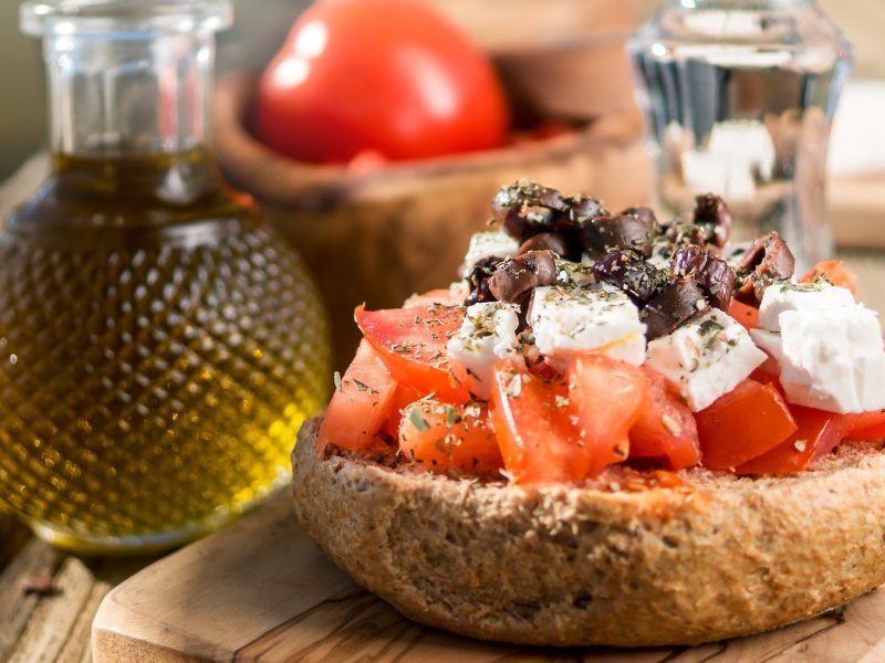 Dakos mit Tomaten, Feta, Oliven und Oregano, daneben eine Flasche mit Olivenöl von vorne fotografiert.