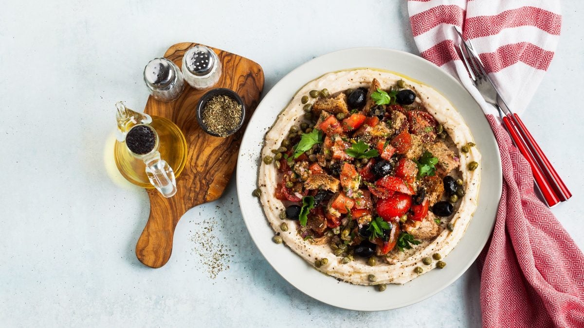 Draufsichat auf Dakos-Salat mit Hummus auf einem Teller, daneben ein Brett mit Öl und Gewürzen.