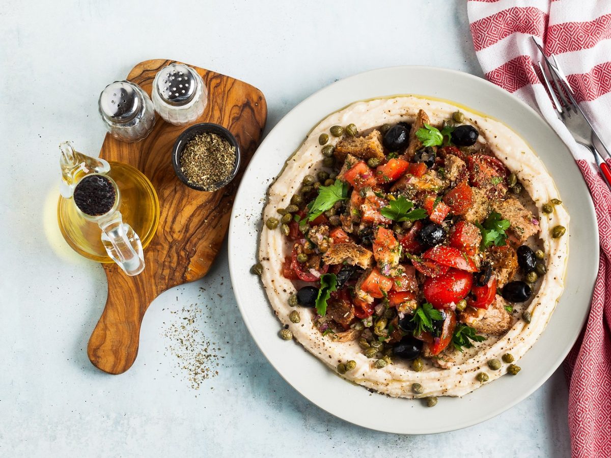 Draufsichat auf Dakos-Salat mit Hummus auf einem Teller, daneben ein Brett mit Öl und Gewürzen.