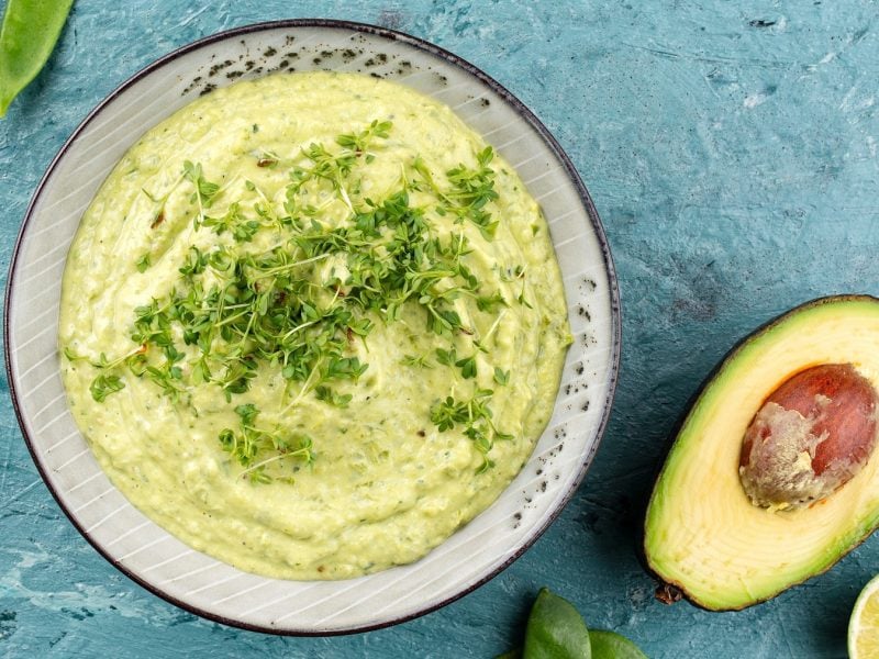 Eine graue Schale mit dem Avocado-Dip auf hellblauem Untergrund direkt von oben fotografiert. Daneben noch eine halbierte Avocado, Limette und ein paar Zuckerschoten.