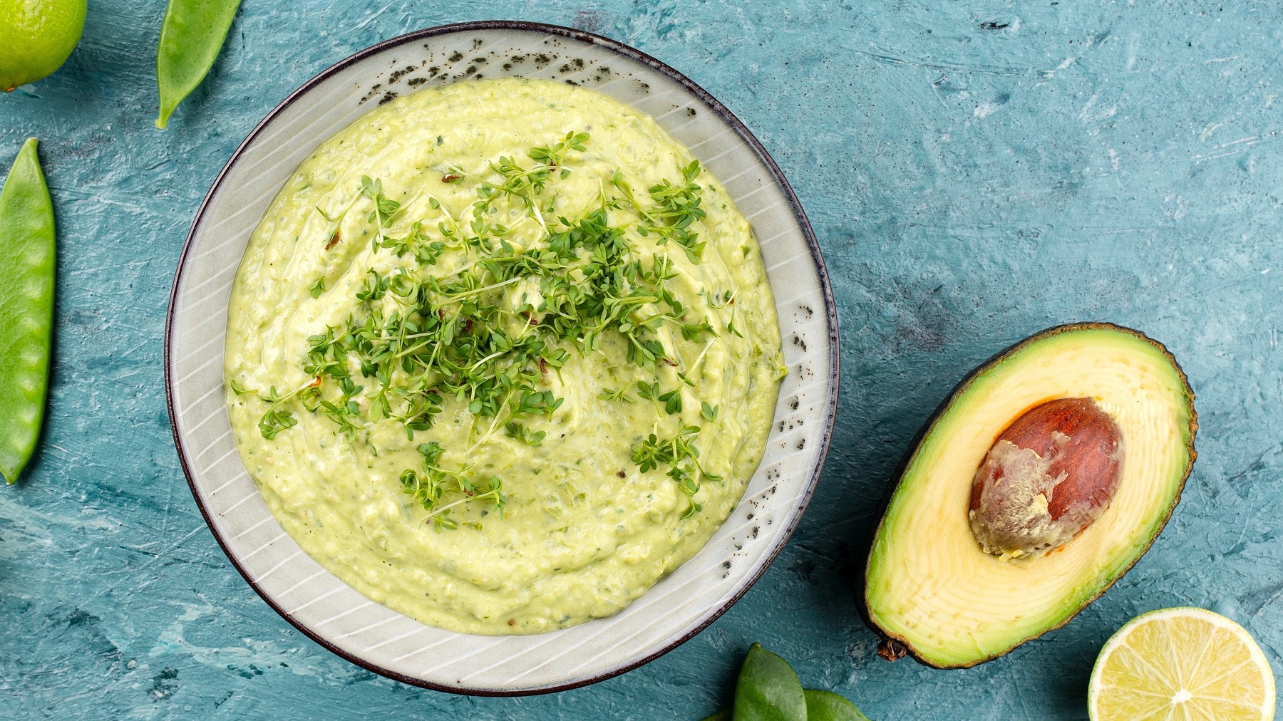 Eine graue Schale mit dem Avocado-Dip auf hellblauem Untergrund direkt von oben fotografiert. Daneben noch eine halbierte Avocado, Limette und ein paar Zuckerschoten.