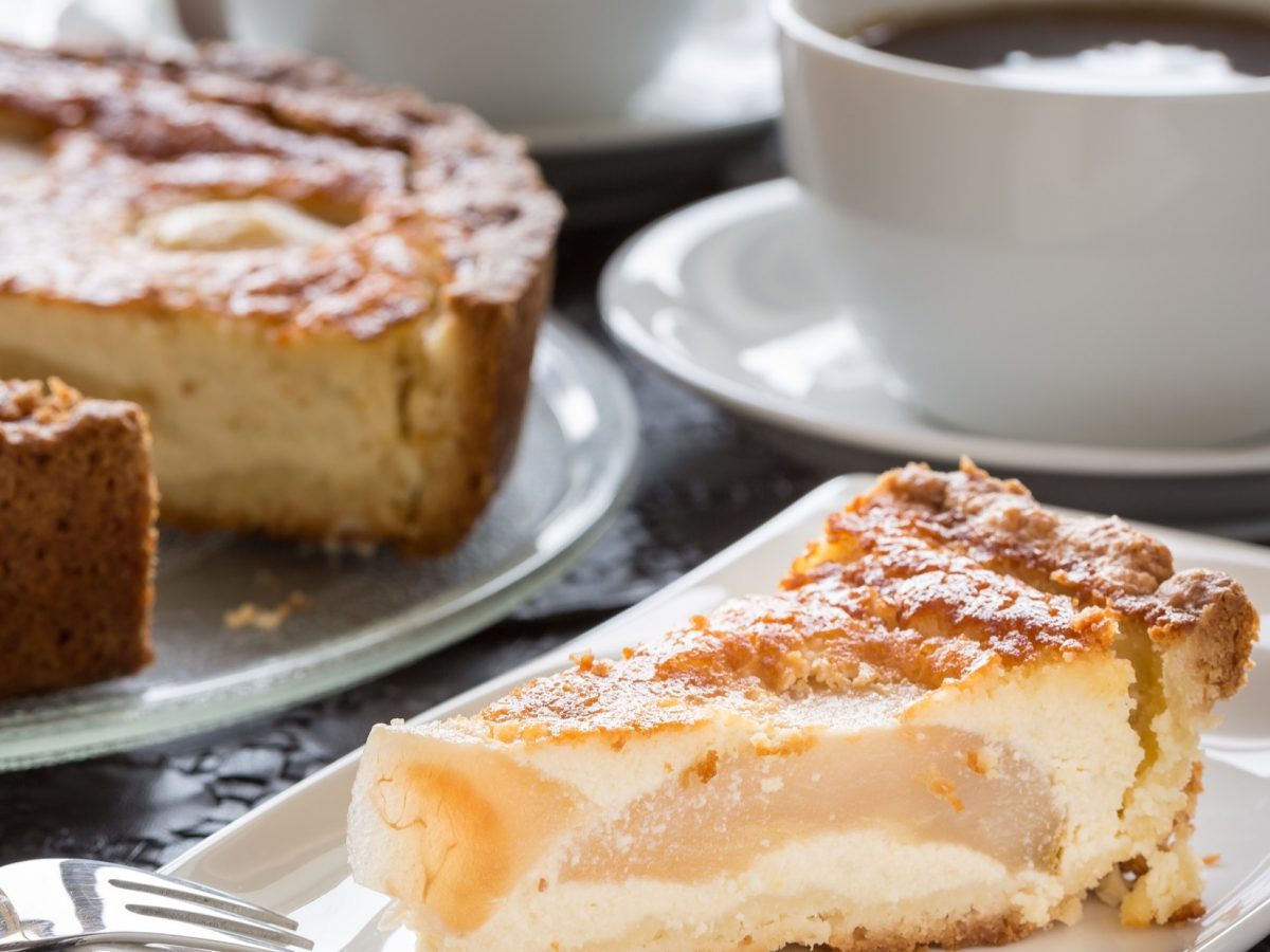 Frontal: Ein gedeckter Tisch mit einem Stück Birnen-Schmandkuchen, einer Tasse Kaffee und einem angeschnittenem Kuchen.