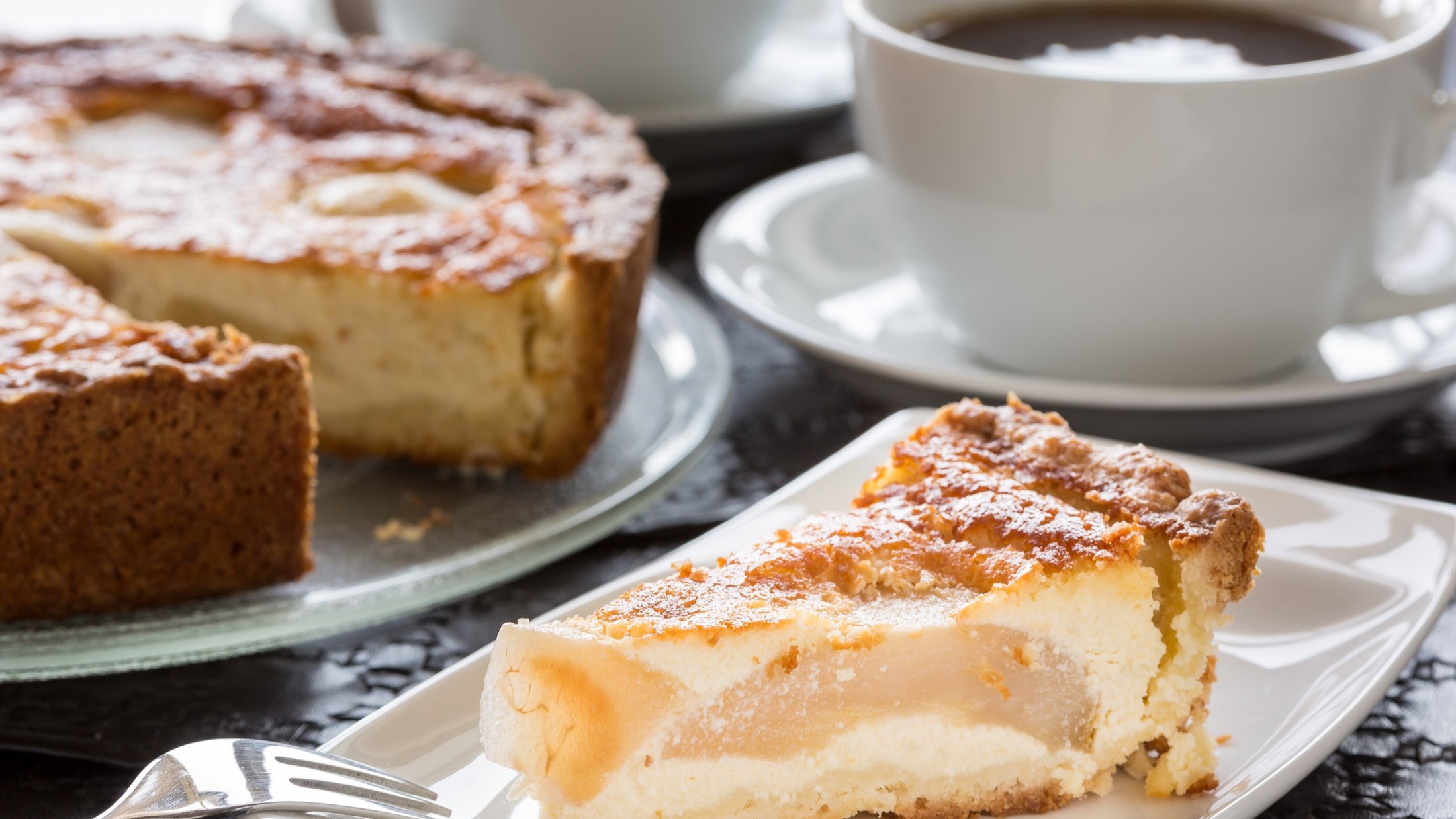 Frontal: Ein gedeckter Tisch mit einem Stück Birnen-Schmandkuchen, einer Tasse Kaffee und einem angeschnittenem Kuchen.