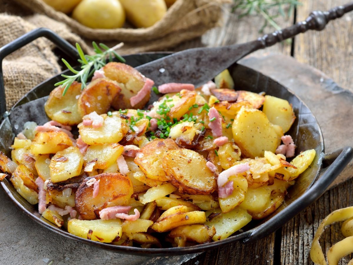 Draufsicht schräg: Eine gusseiserne Pfanne Knusprige Bratkartoffeln aus rohen Kartoffeln mit Zwiebeln und Speck auf einem Untergrund aus Holz. Daneben liegen Kartoffelschalen.