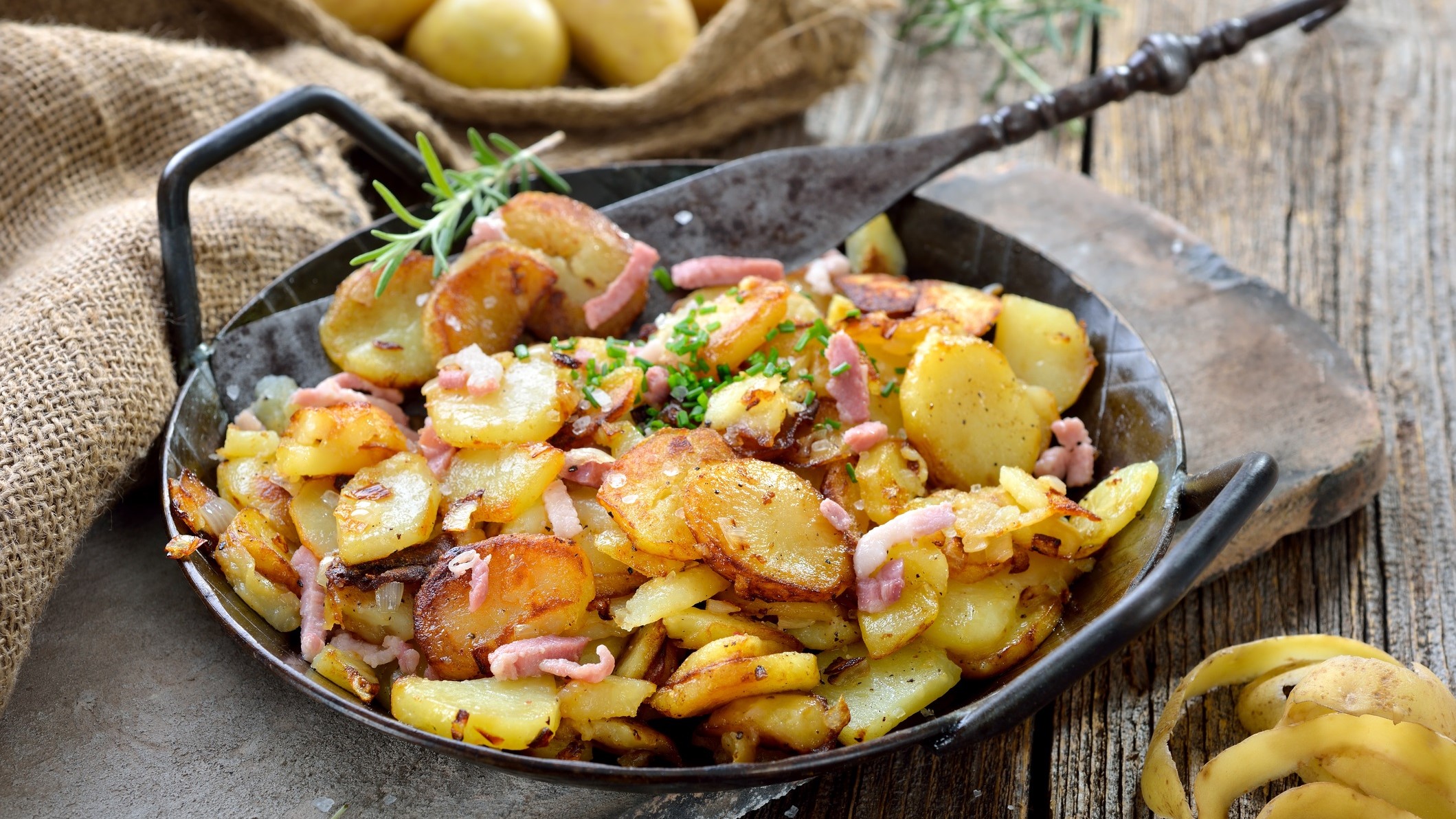 Draufsicht schräg: Eine gusseiserne Pfanne Knusprige Bratkartoffeln aus rohen Kartoffeln mit Zwiebeln und Speck auf einem Untergrund aus Holz. Daneben liegen Kartoffelschalen.